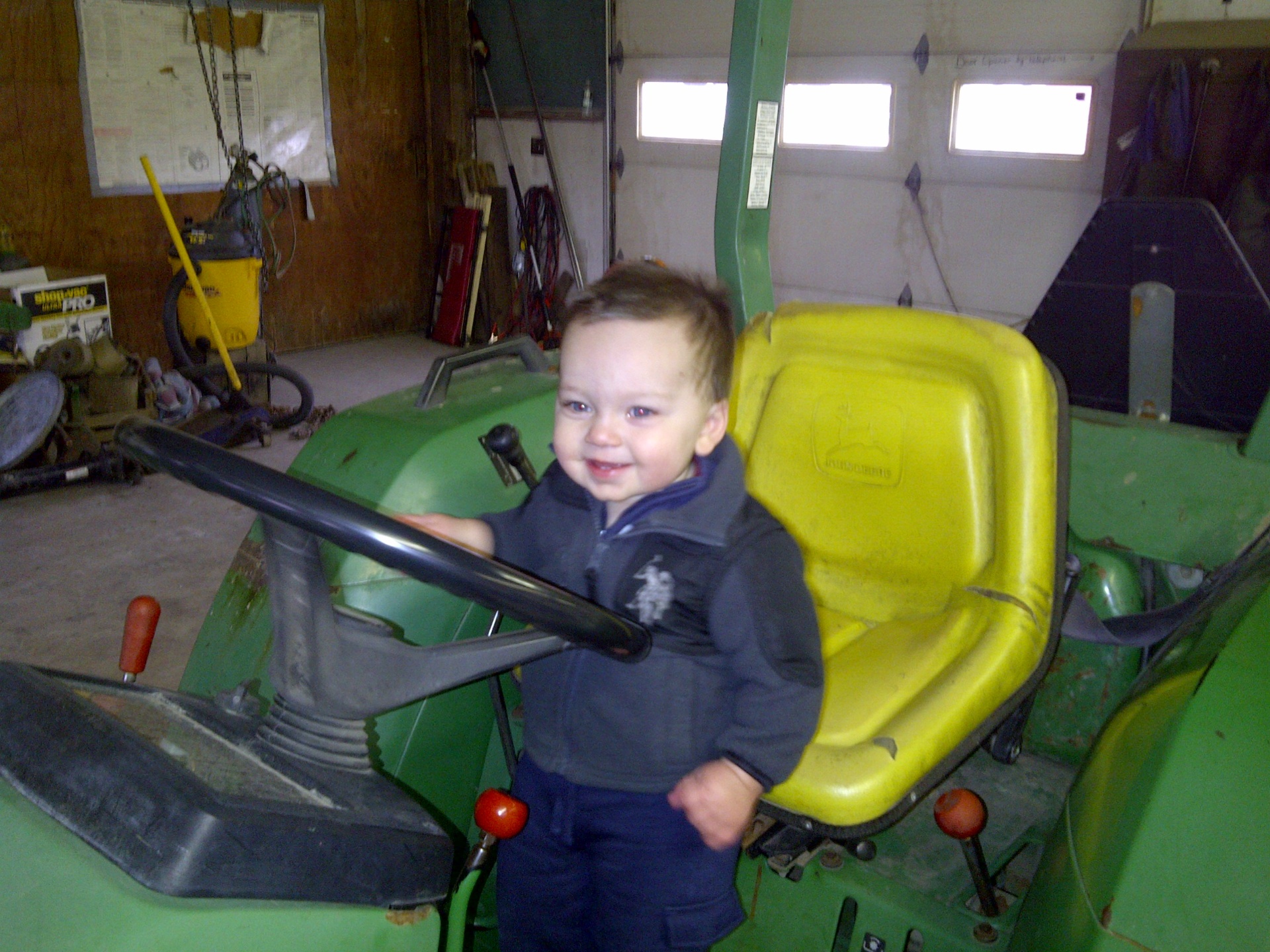 Toddler Carter standing on a John Deere tractor in Grandpa's shop